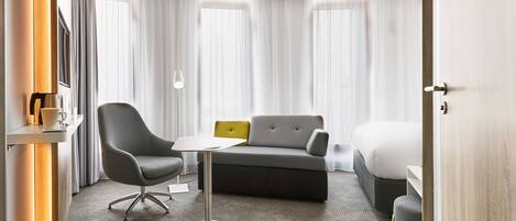 Moderni hotellihuone, jossa on harmaa sohva, pieni pyöreä pöytä, harmaa nojatuoli ja sänky.