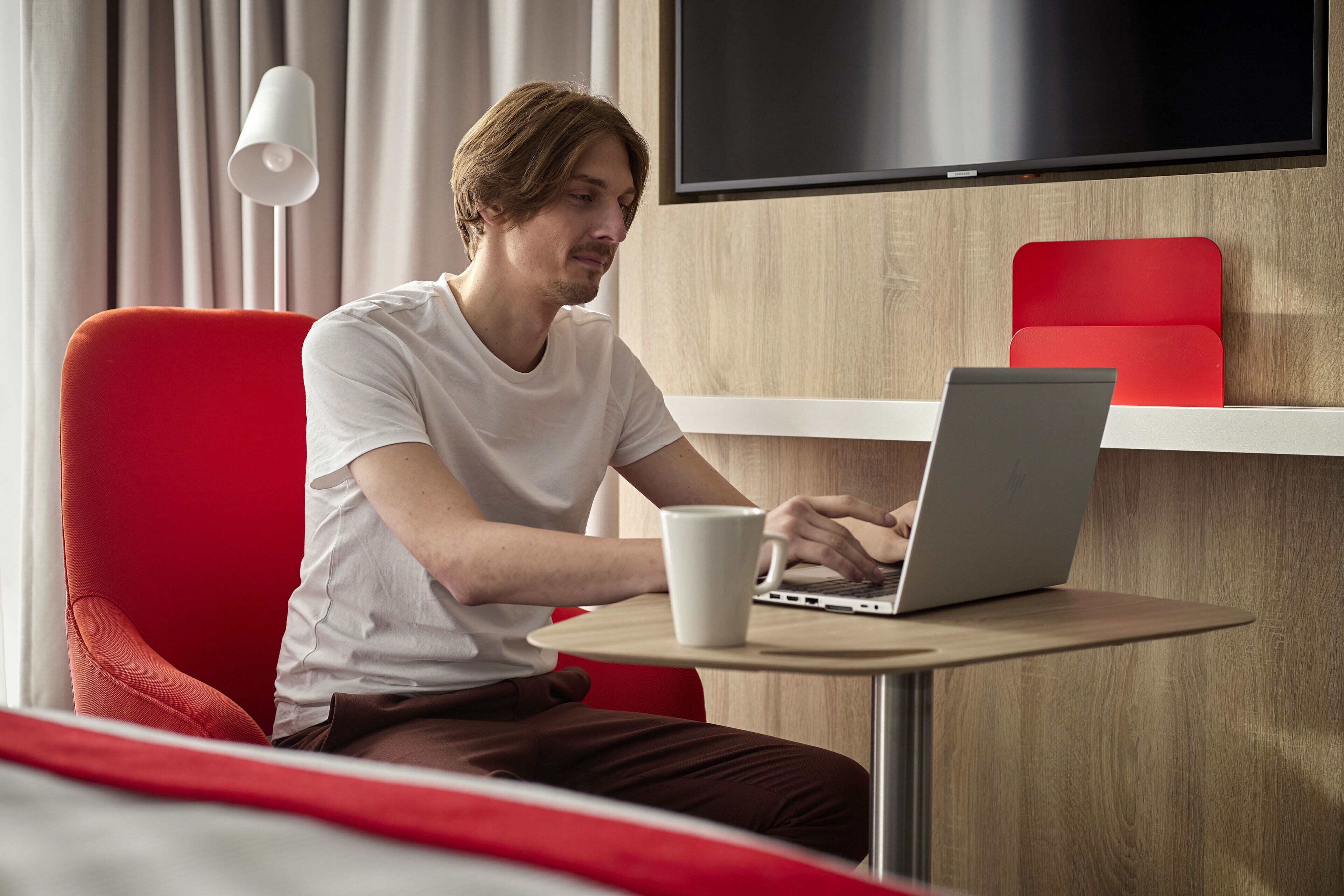 A person seated at a round table with a laptop and a white mug, in a room with a red chair and a TV mounted on the wall.