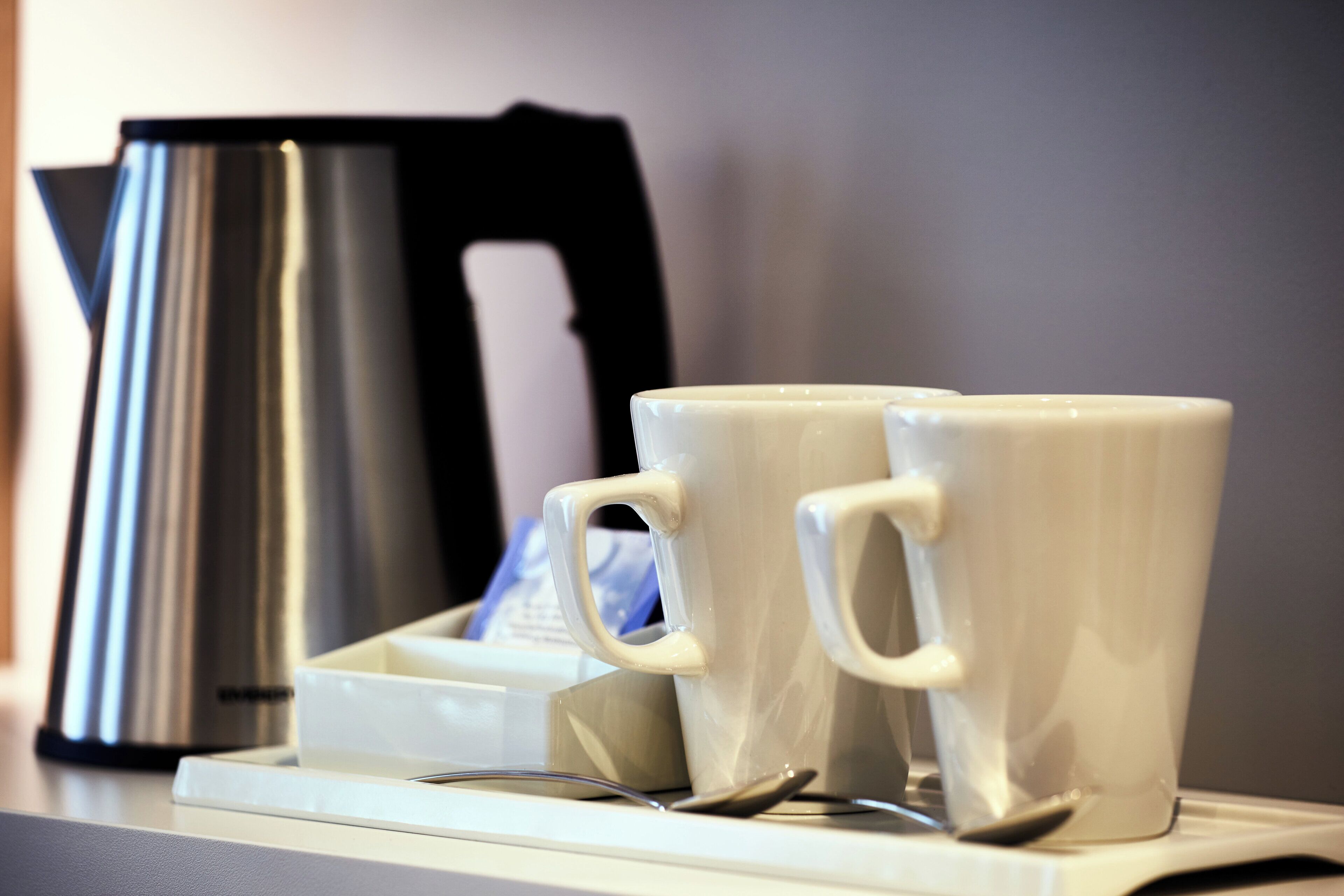 Two white ceramic mugs on a tray with a spoon, next to a stainless steel kettle.