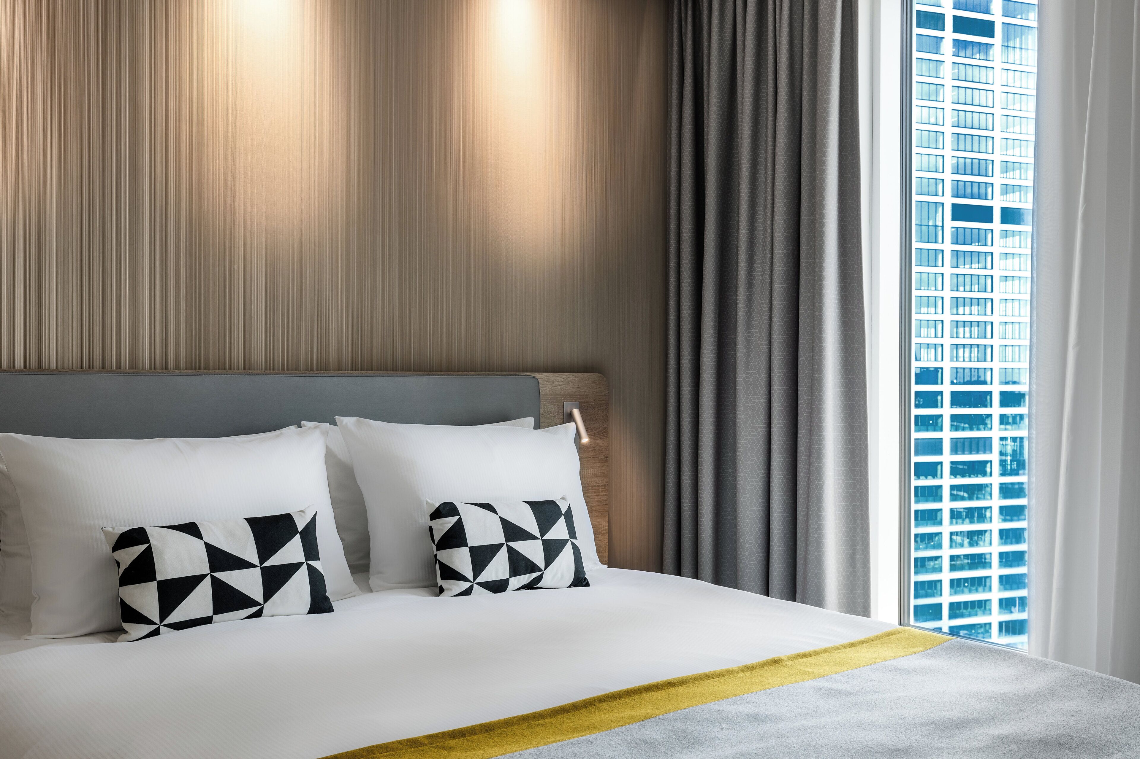 A hotel room with a large bed, two pillows with a geometric pattern, a wooden headboard, and a view of a cityscape through a window with grey curtains.