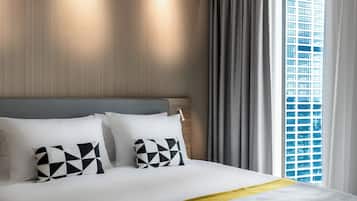 A hotel room with a large bed, two pillows with a geometric pattern, a wooden headboard, and a view of a cityscape through a window with grey curtains.