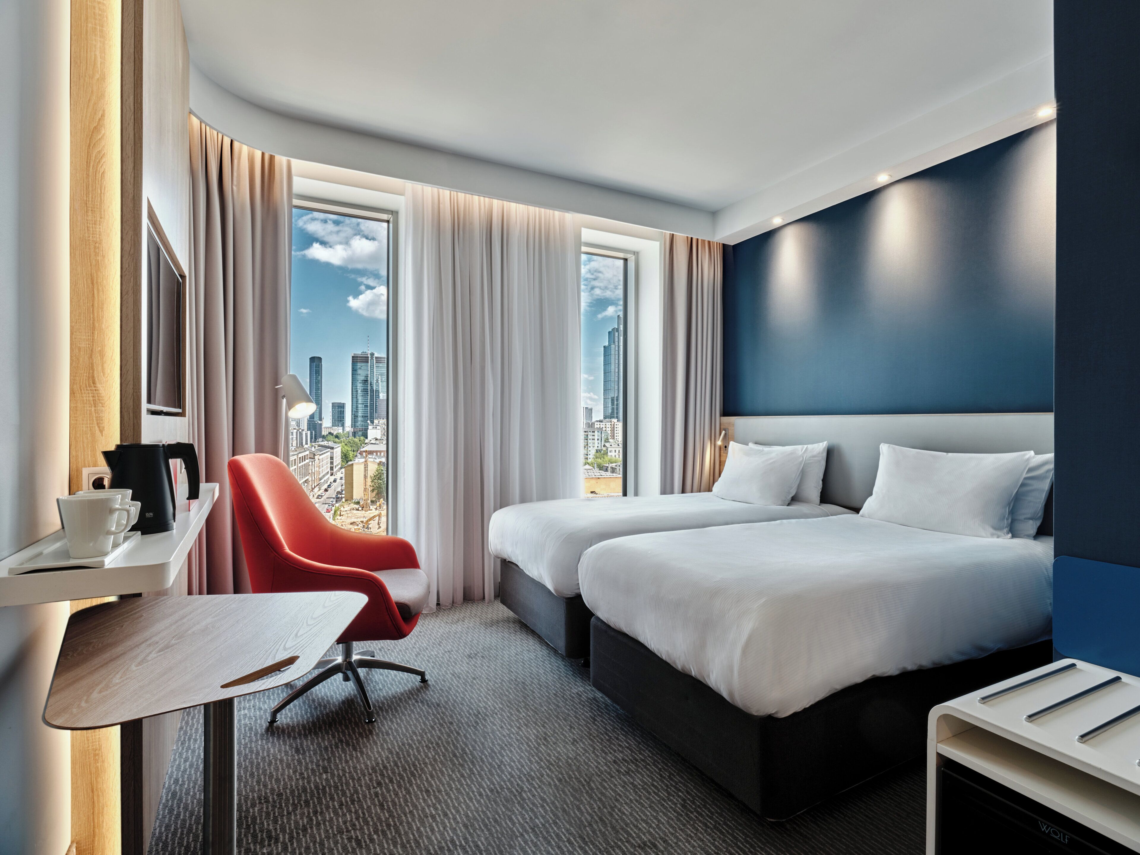 A hotel room with a city view, a desk with a coffee maker, a red chair, and a bed with white linens.