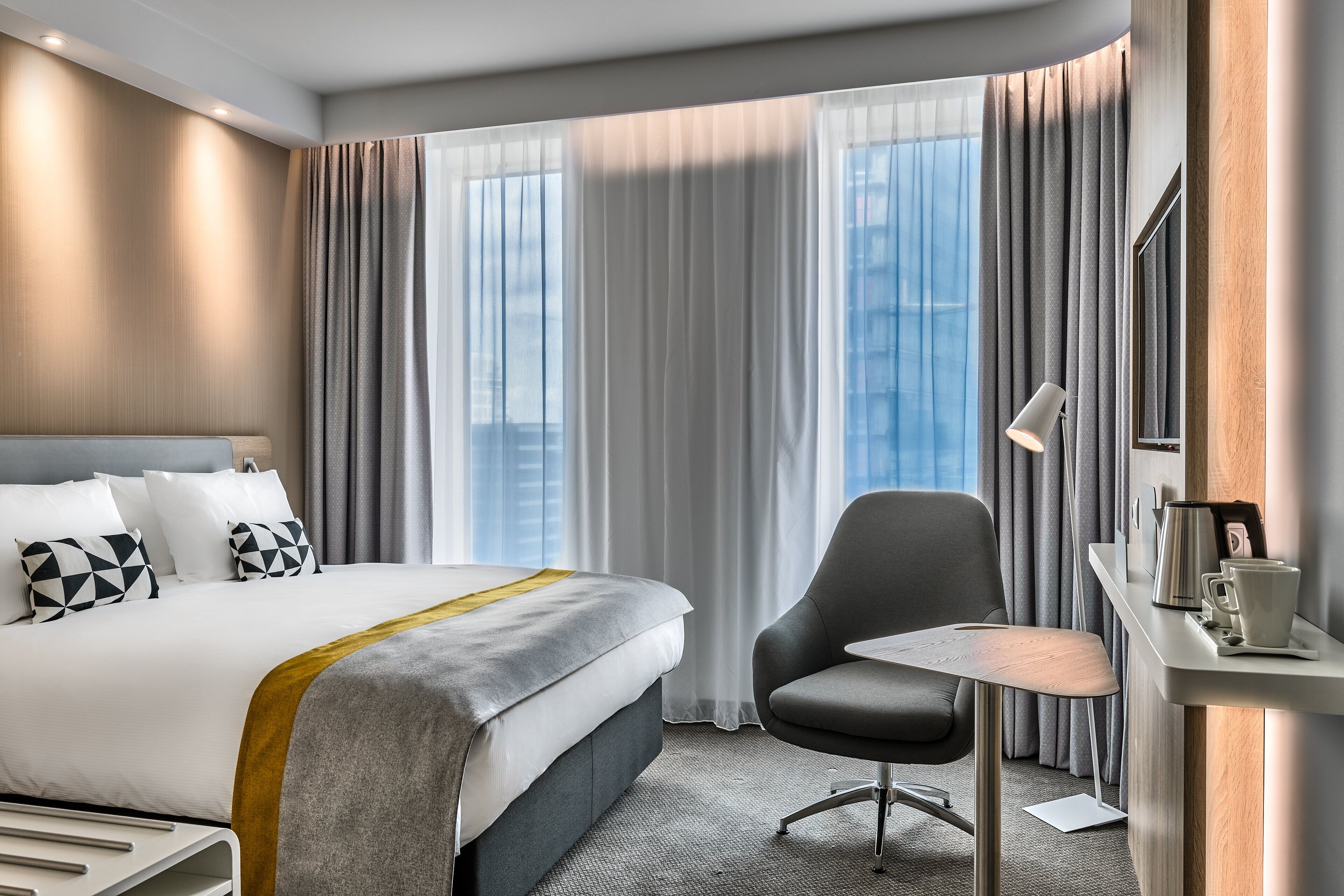 Ein modernes Hotelzimmer mit einem großen Bett, einem Schreibtisch mit Stuhl und Blick auf die Stadt durch große Fenster.