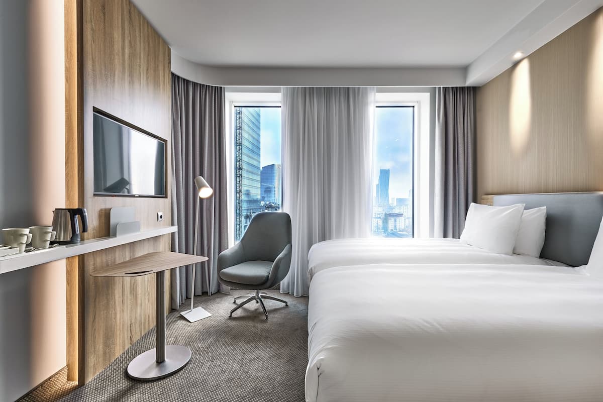 A hotel room with a large bed, a desk with a chair, a TV, and a view of the cityscape through large windows.