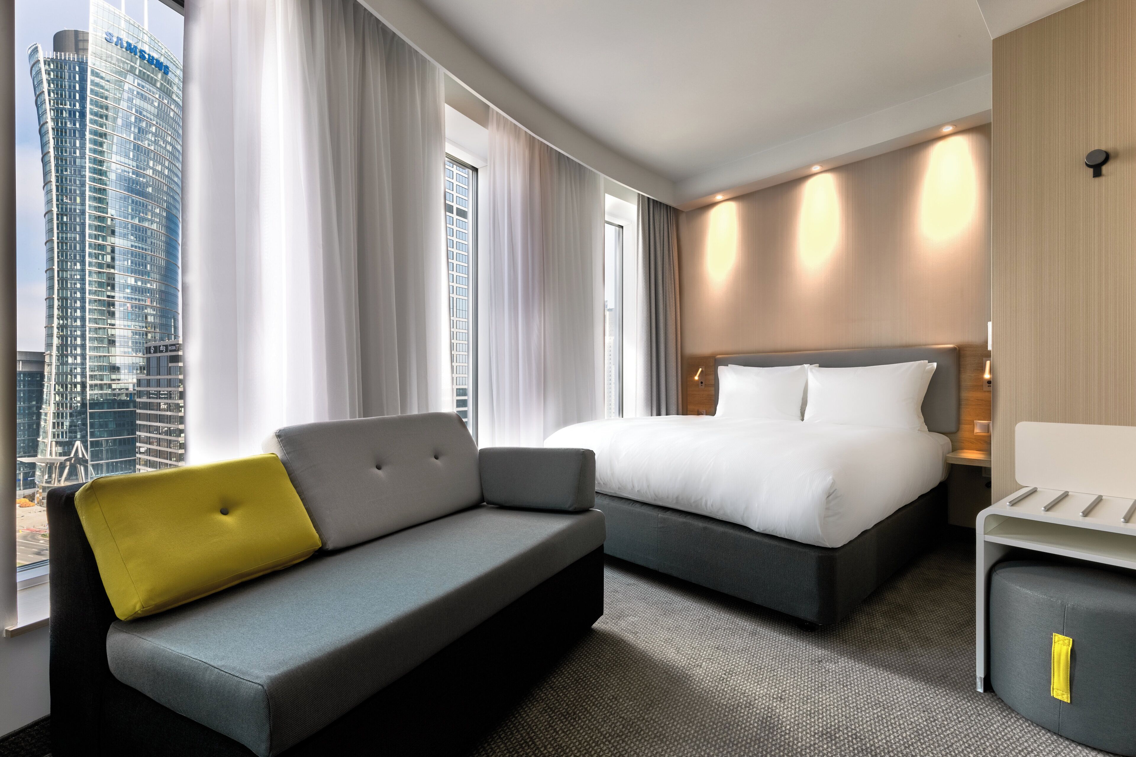 A hotel room with a bed, a sofa, a small table, and a view of a cityscape.