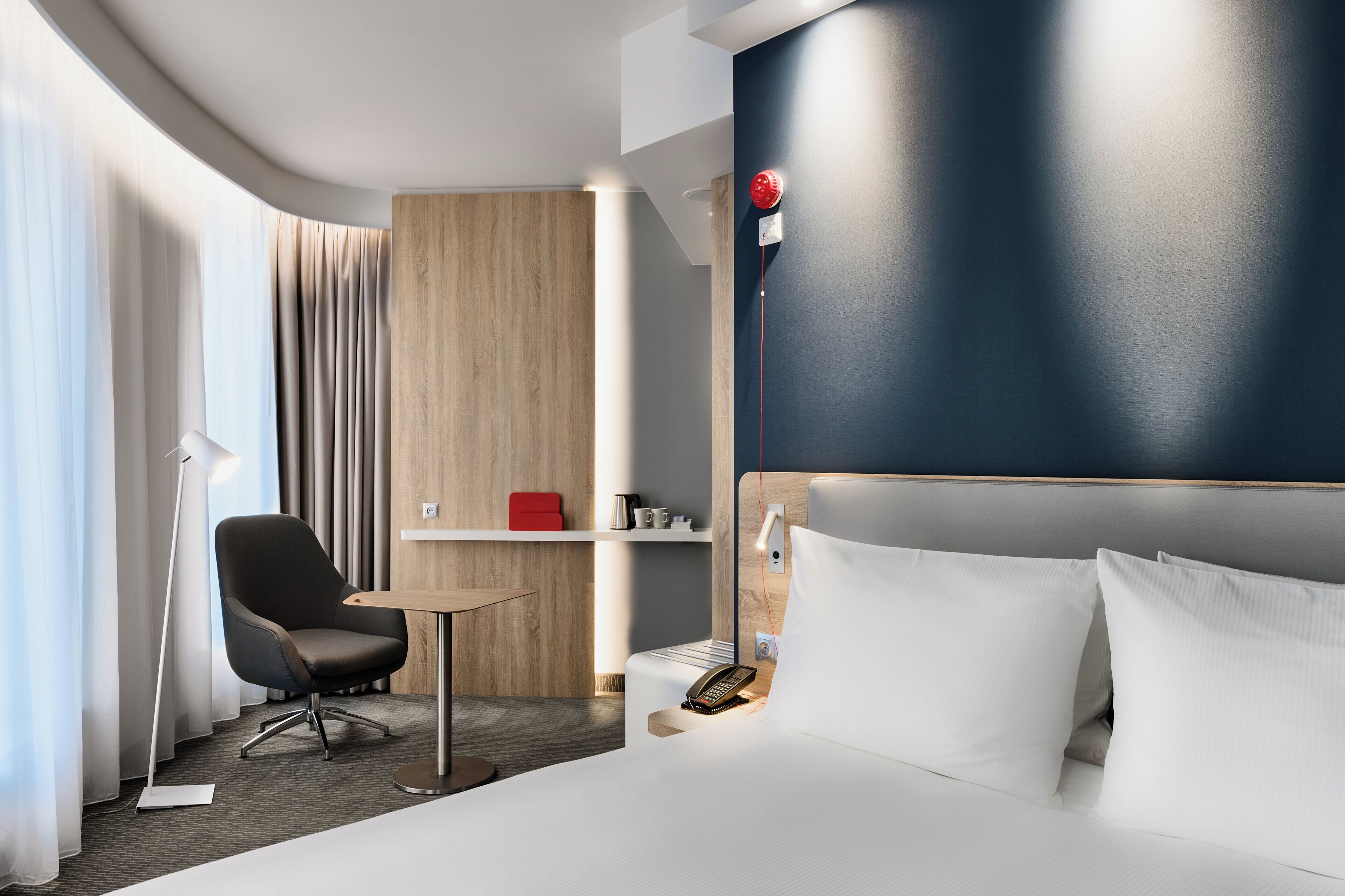 Habitación de hotel con cama, escritorio, silla, mesita de noche, teléfono y panel de control en la pared.