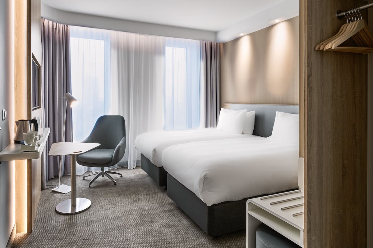 A hotel room with a large bed, a desk with a chair, a small table, and a window with curtains.