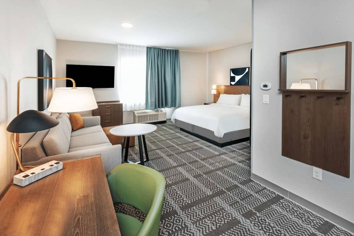 A hotel room with a bed, a sofa, a desk, a chair, a TV, and a mirror.