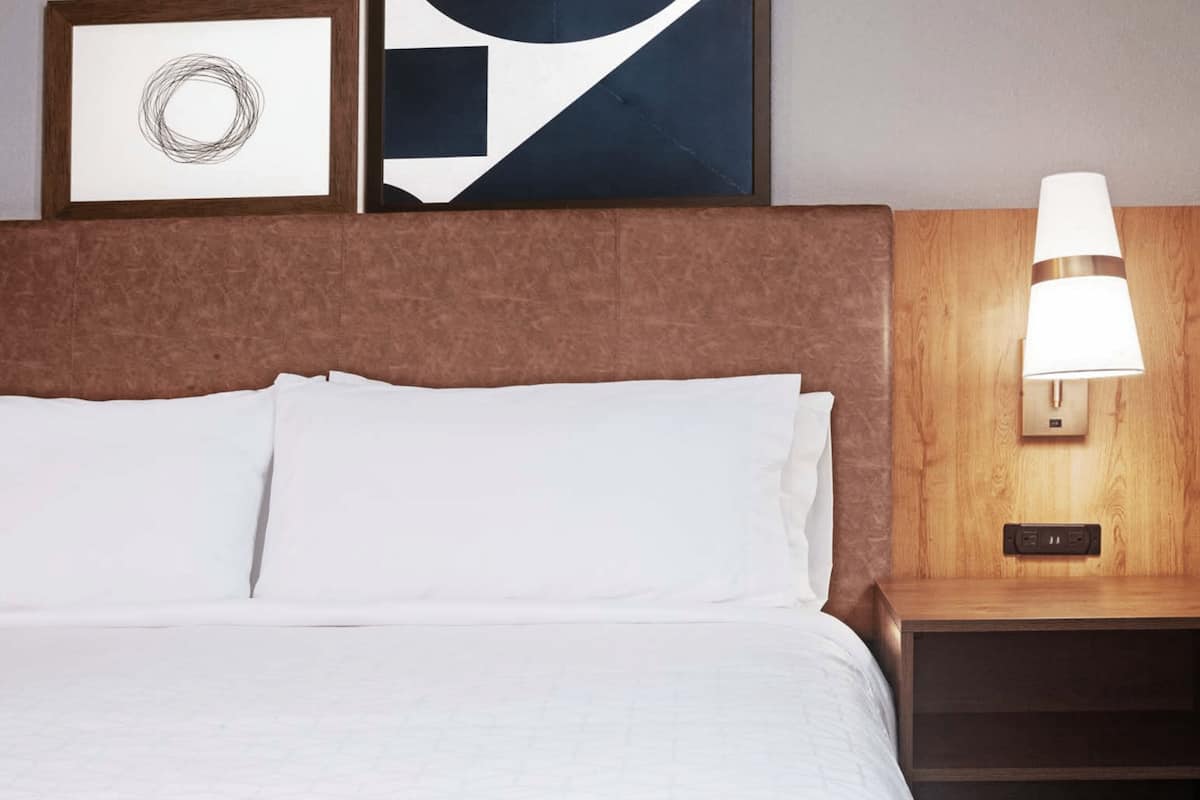 A hotel room with a large bed, a wooden headboard, a nightstand with a lamp, and two framed artworks on the wall.