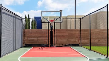 Un terrain de basketball en plein air avec une surface de jeu rouge, entouré d’une clôture en treillis noir, et une clôture en bois en arrière-plan.