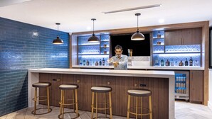 A modern bar area with a wooden bar counter, three stools, pendant lighting, and a shelf with bottles. - Staybridge Suites Waco South - Woodway by IHG (Waco)