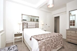 Ein Schlafzimmer mit einem großen Bett, einem Nachttisch, einer Kommode, einem Spiegel und einer Tür zu einem anderen Raum.