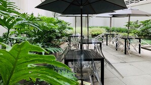 Ein Terrassenbereich im Freien mit mehreren Tischen und Stühlen, umgeben von üppigen Pflanzen und Sonnenschirmen, die Schatten spenden.