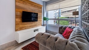 Un soggiorno moderno con una TV a schermo piatto montata su una parete rivestita in legno, un divano grigio con un cuscino rosso e un'ampia finestra vista edifici.