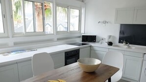 A modern kitchen with white cabinetry, a wooden dining table, and a large window with a view of palm trees. - Appartement de charme , Marina Royale, Marigot (Marigot)