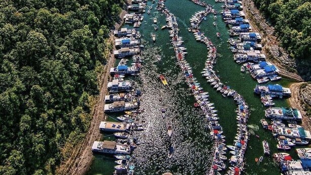 Une vue aérienne d’une rivière où de nombreux bateaux sont amarrés le long des rives, le tout étant entouré d’une forêt dense.