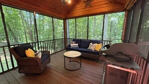 Une véranda couverte avec un plafond en bois, un ventilateur de plafond, un barbecue encastré, des meubles en osier et une vue sur une forêt.