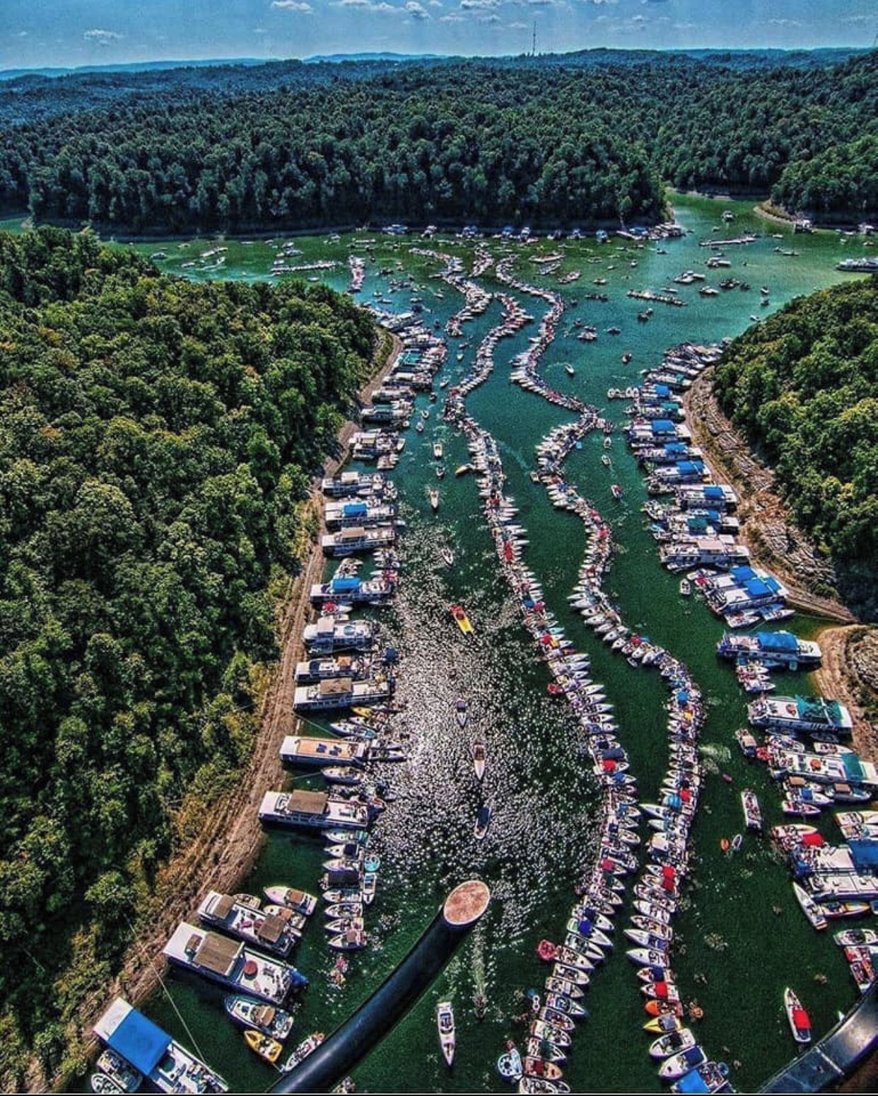 An aerial view of a river with numerous boats docked along the banks, surrounded by dense forest.