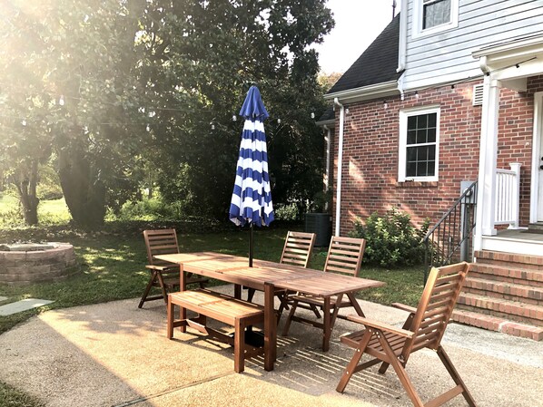 A patio with a wooden table and chairs, a blue and white striped umbrella, and a brick house in the background. - Waterfront Retreat with Office and Private Dock, Boats! (Weems)