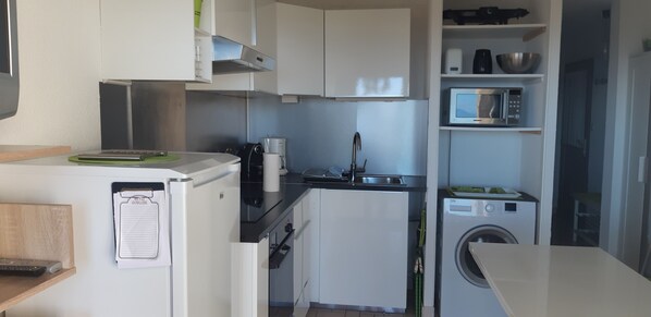 A compact kitchen with white cabinets, a microwave, a coffee maker, and a washing machine. - One-bedroom apartment with ocean view and access 30 meters away (Sarzeau)