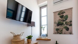 A modern bedroom with a wooden dresser, a flat-screen TV, a window with a view, a wall-mounted air conditioner, and a botanical tapestry. - AZUR - LES COCONS - Studio Apartment, Sleeps 2 (Marseille)