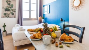 A hotel room with a bed, a dining table with breakfast items, a window with curtains, and a wall-mounted mirror. - AZUR - LES COCONS - Studio Apartment, Sleeps 2 (Marseille)