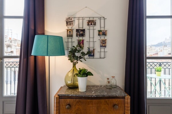Ein Zimmer mit einem Holzkasten, einer türkisfarbenen Lampe, einer Vase mit Grünpflanzen und einer an der Wand befestigten Fotocollage.