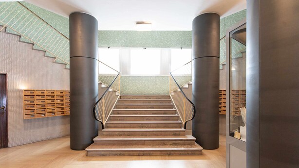 Una escalera moderna con barandillas de metal, flanqueada por grandes columnas cilíndricas, que lleva a un rellano con pared estampada y panel de madera.