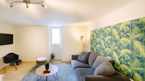 A modern living room with a grey sofa, a round coffee table, a black chair, a small potted plant, and a wall with a tropical leaf pattern.