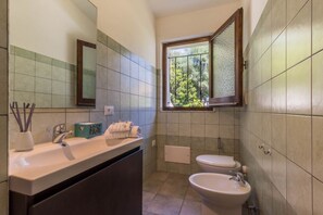Un baño con lavamanos, dos inodoros, una ventana que da a zonas verdes y un espejo.