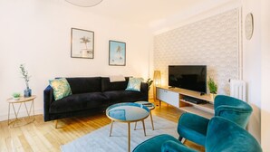 A modern living room with a dark sofa, teal armchairs, a round coffee table, and a flat-screen TV. - L'Imperti'Nantes- T3bis de Standing (Nantes)