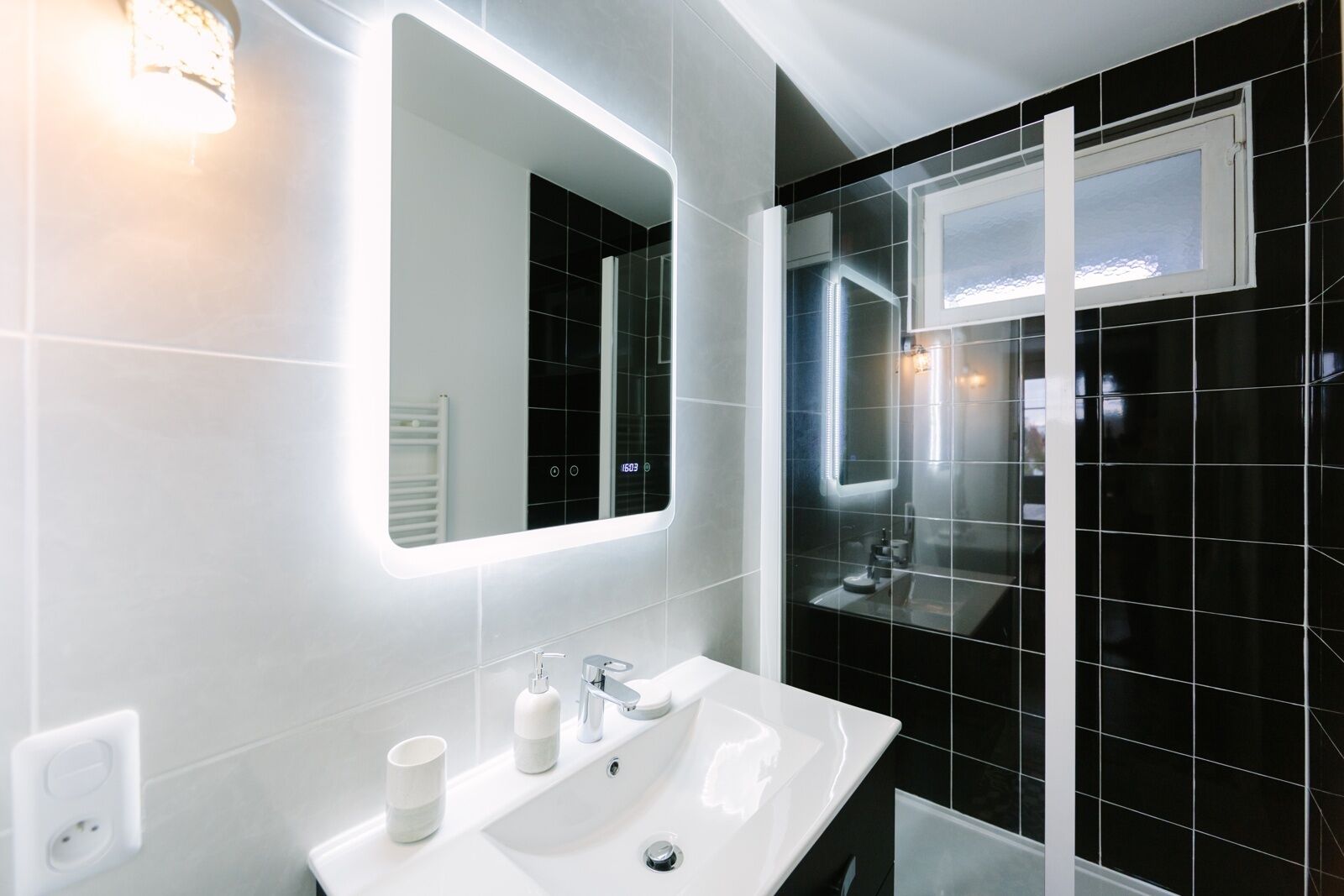 Een moderne badkamer met een grote spiegel, een wit wastafelmeubel en zwart betegelde wanden.