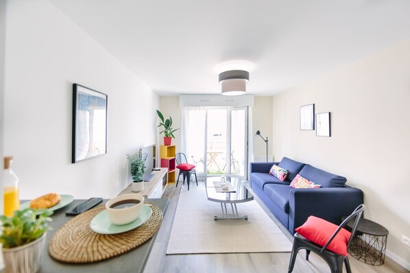 A modern living room with a blue sofa, a coffee table, and a dining area with a plant and a bottle of orange juice.