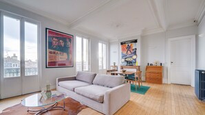 A spacious living room with a large window, a glass coffee table, a sofa, and a poster of a movie on the wall. - LE VICTOR - Grand et lumineux T3 (Bordeaux)