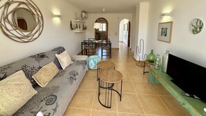 A living room with a sofa, a coffee table, and a TV. There is a dining area with chairs and a table in the background. - Apartamento Jardines PG004 - Benitachell, Costa Blanca (Benitachell)