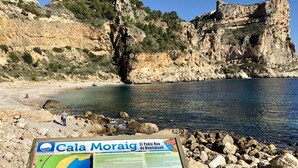 A rocky beach with a signboard displaying information about Cala Moraig. - Apartamento Jardines PG004 - Benitachell, Costa Blanca (Benitachell)