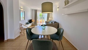A modern living room with a round dining table, two chairs, a sofa, and a hanging light fixture. - Apartamento Jardines PG010 - Benitachell, Costa Blanca (Benitachell)