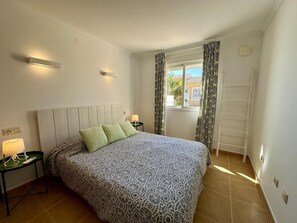 Ein Schlafzimmer mit einem Bett, zwei Nachttischlampen, einem Fenster mit Vorhängen und einer an der Wand stehenden Leiter.