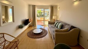A living room with a sofa, TV, and a small table. There is a balcony with a chair and a table outside. - Apartamento Jardines PG010 - Benitachell, Costa Blanca (Benitachell)