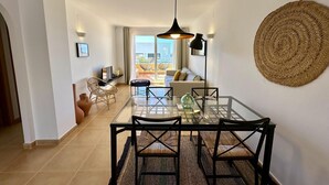 Ein moderner Essbereich mit einem Glastisch und schwarzen Stühlen, einem gewebten Wandbehang und Blick ins Freie durch eine Schiebetür.