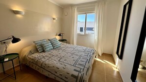 Ein Schlafzimmer mit einem Bett, zwei Nachttischen, einem Fenster mit Vorhängen und einem Wandspiegel.