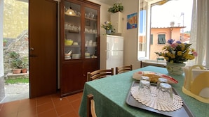 A dining area with a table set for two, a cabinet with glass doors, a refrigerator, and a window with a view of buildings. - The house in the carob trees with breathtaking views (Arcola)