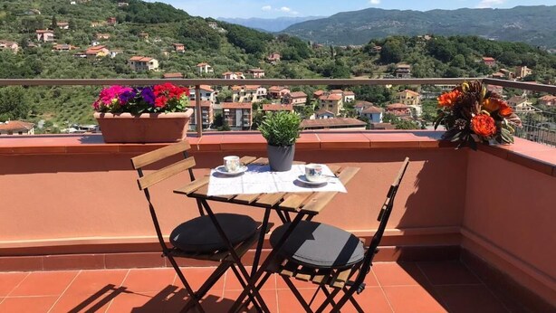 A terrace with a table set for two, a potted plant, and a view of a hillside village. - The house in the carob trees with breathtaking views (Arcola)