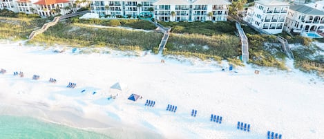 Un hotel frente a la playa con varias construcciones, una playa arenosa con sillas azules y una vista despejada del océano.