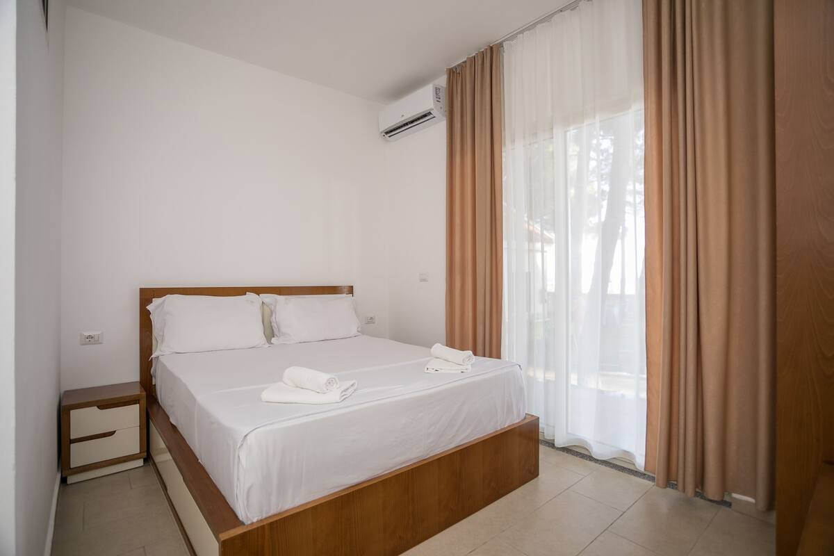 Una cama bien tendida con ropa blanca y un cabecero de madera, una mesita de noche con cajón y una ventana con cortinas traslúcidas.
