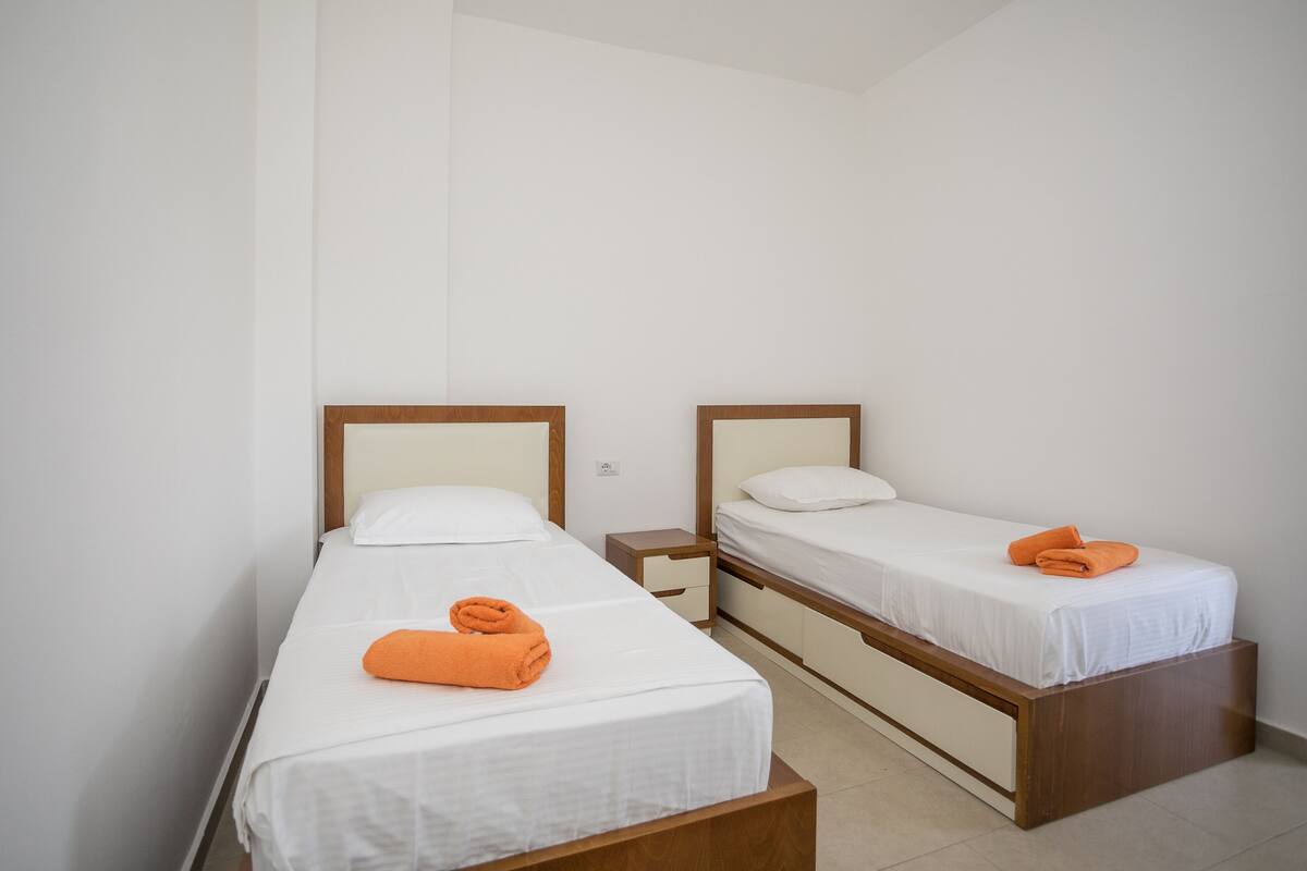 Una habitación pequeña y limpia con dos camas individuales, cada una con cabecero de madera y ropa de cama blanca. Hay una mesita de noche con cajón entre las camas.