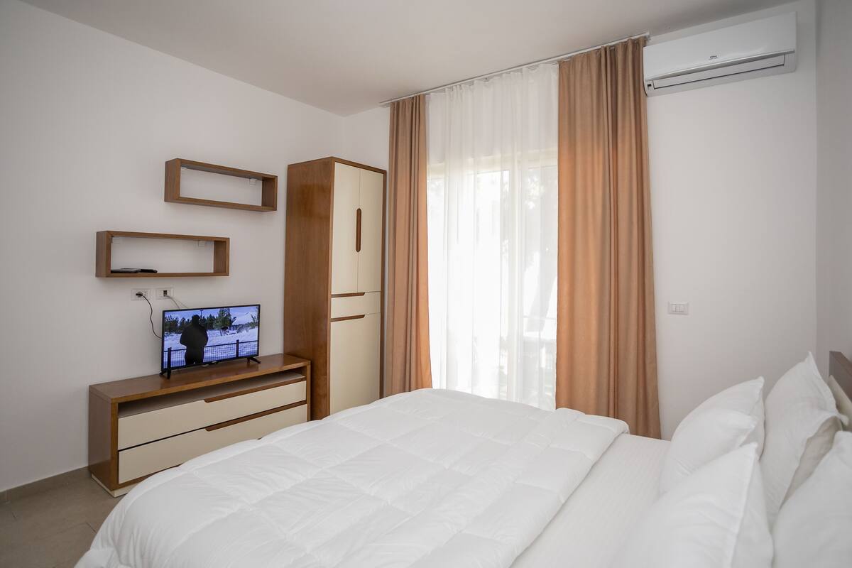 Habitación de hotel con cama, televisor sobre una consola de madera, armario y ventana con cortinas.