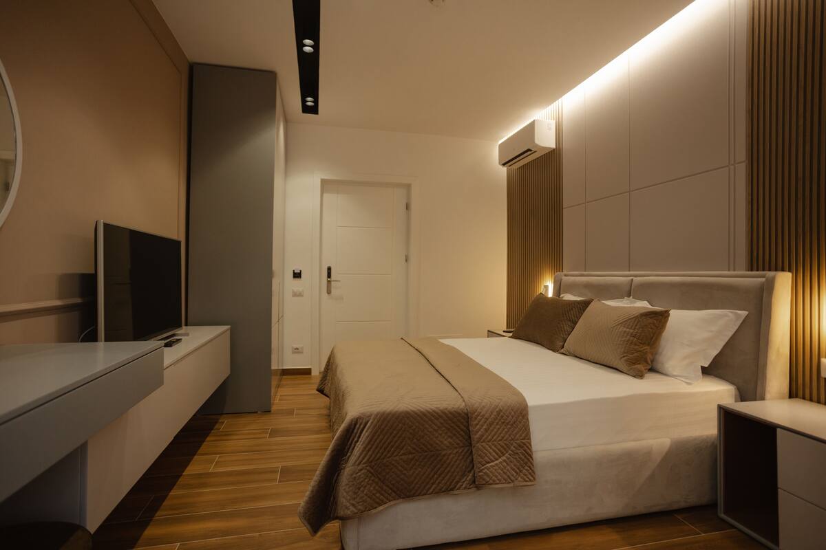 Un dormitorio moderno con una cama grande, mesitas de noche, un televisor y una puerta que da a otra habitación.