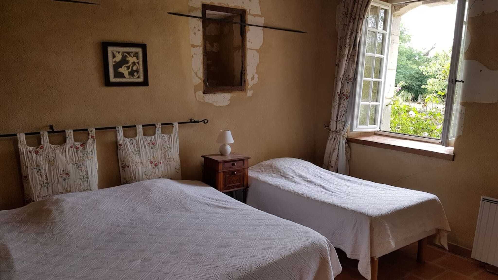 Une chambre avec deux lits, une petite table de chevet avec une lampe, un tableau encadré au mur et une fenêtre avec des rideaux.