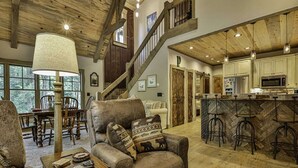 A rustic interior with a wooden ceiling, a bar area with stools, a sofa with patterned cushions, and a dining area with chairs and a table.